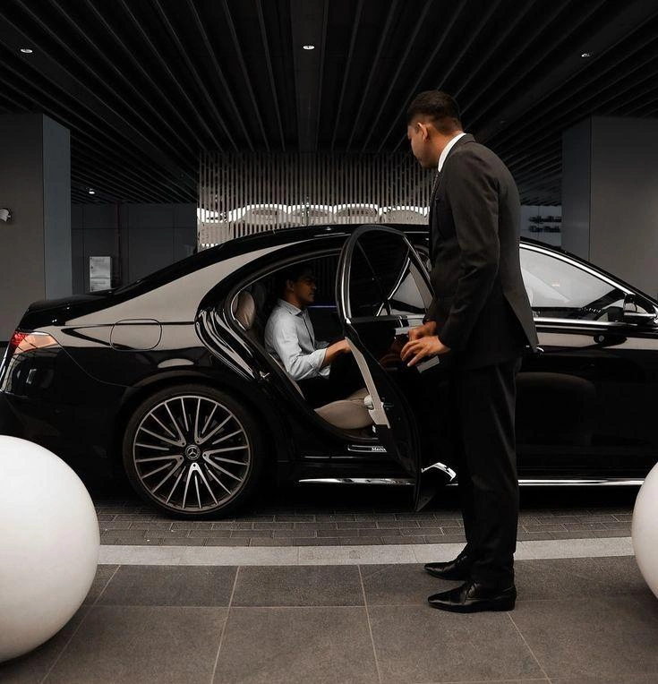 Professional chauffeur assisting a passenger into a black luxury Mercedes-Benz saloon outside a modern building.